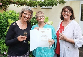 Jubilarin zwischen den Leitungskräften beim Anstoßen und mit der Urkunde in der Hand. 